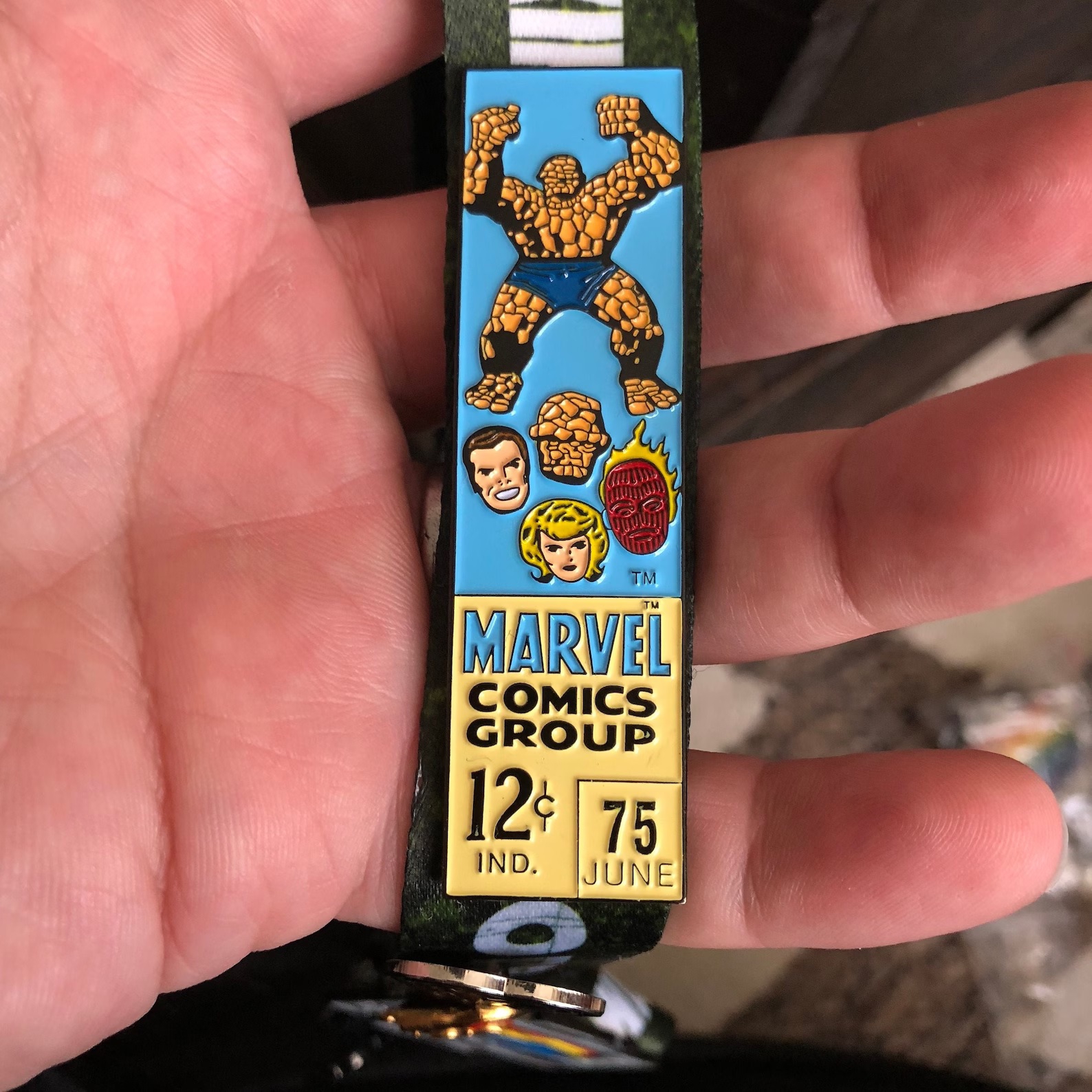 A pin shaped like an old-fashioned price box from a Fantastic Four comic, including the heads of each of the FF and a full-sized Thing above them