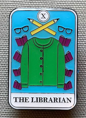 an enamel pin of a sweater, pencils, glasses, and books that says The Librarian