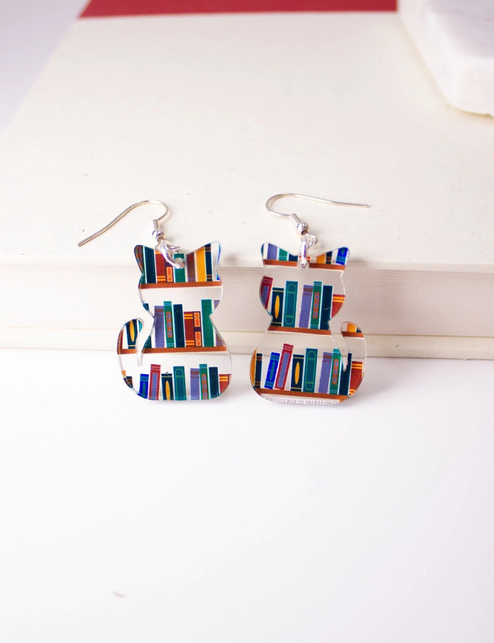 a photo of two earring made of clear plastic in the shape of two cats. Illustrations of colorful books run across each cat-shaped earring.