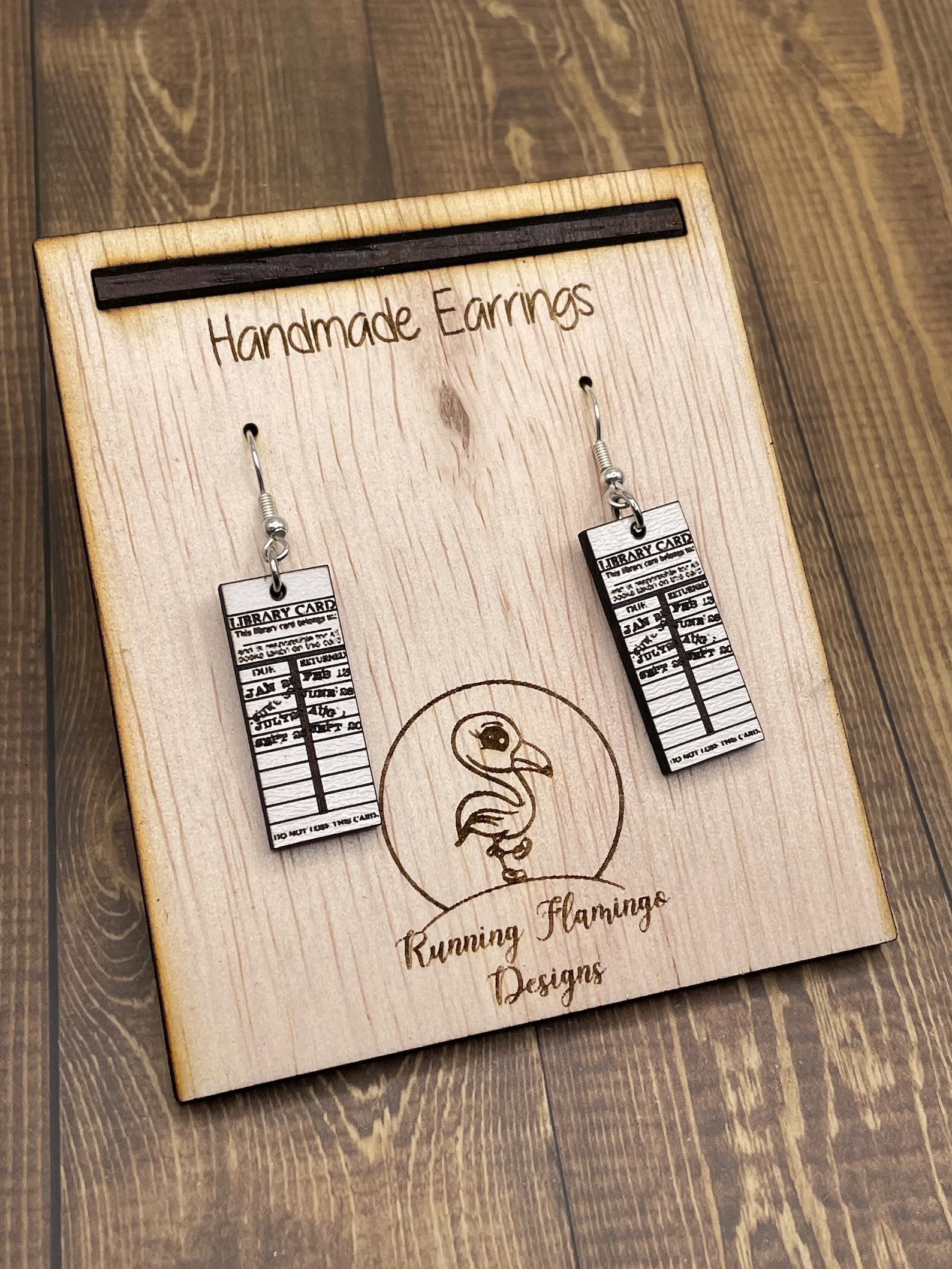a photo of a pair of earring made out of wood. They are made to looking like little library cards stamped with various past due dates.