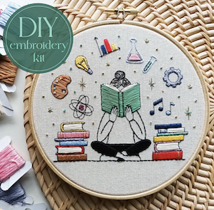 an embroidery of a person sitting down reading with stacks of books and symbols for art, idea, science above their head