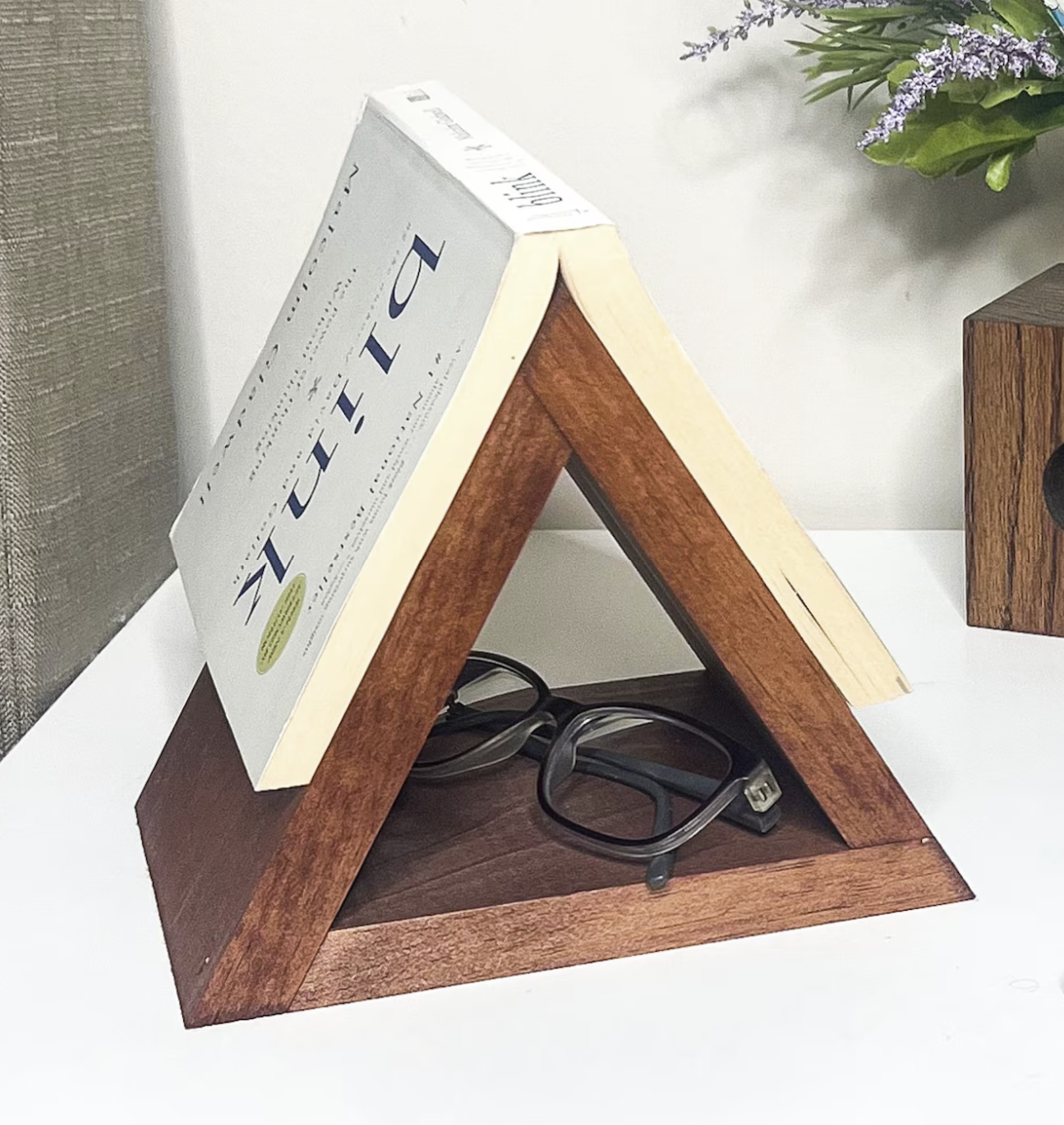 Picture of a triangular wooden book holder on a nightstand with an open paperback book laying on top to hold its spot. 