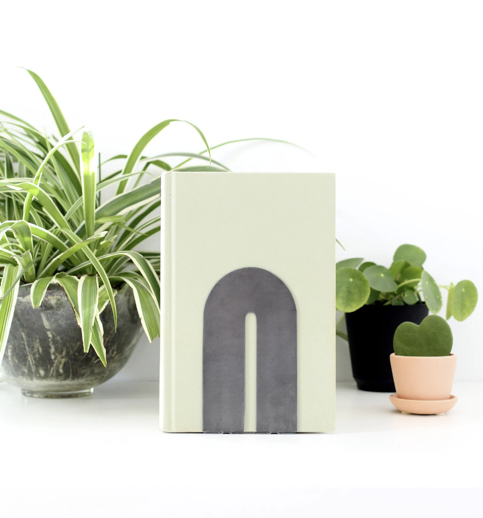 Picture of a light green book being held up with a silver metal arch bookend, surrounded by plants on a white background.