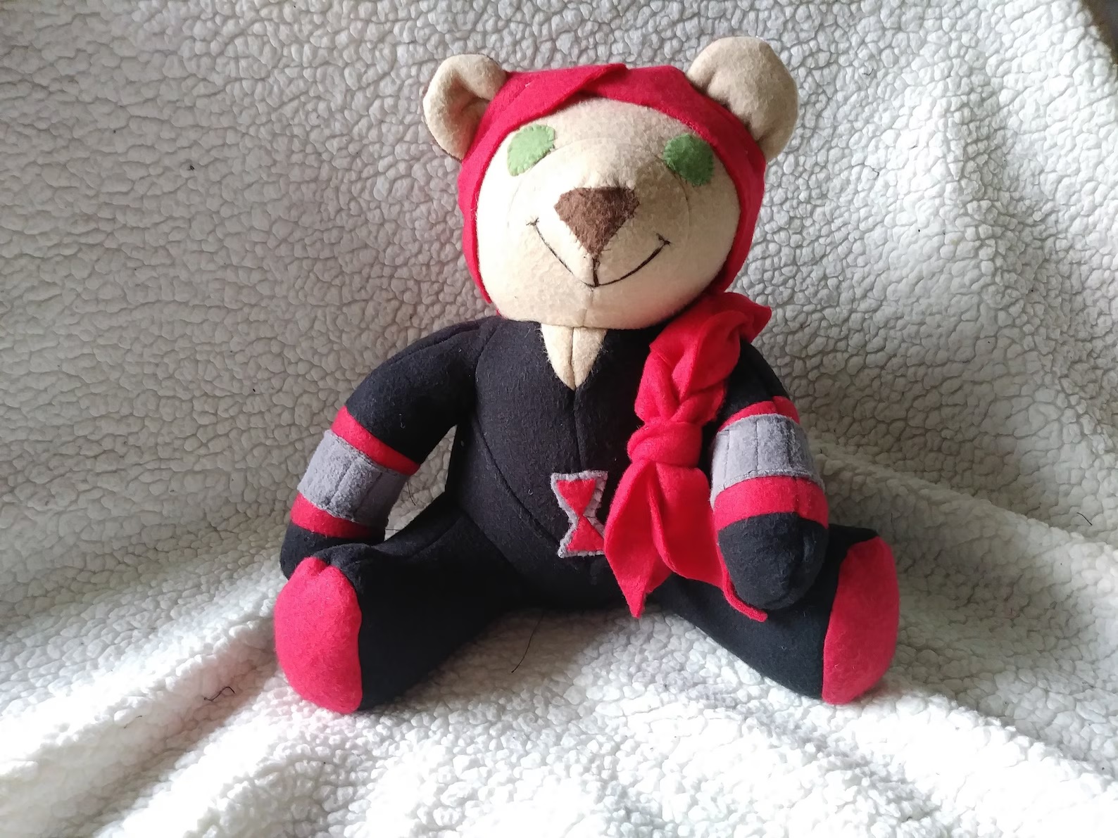 A handmade, off-white teddy bear wearing Black Widow's costume and a red braided cloth for hair. It has green felt eyes and is smiling.