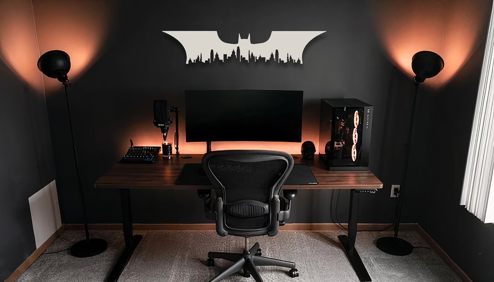A sharp silver Bat-symbol with the silhouette of Gotham City carved into the bottom. It hangs on a black wall above a desk with a monitor on it.