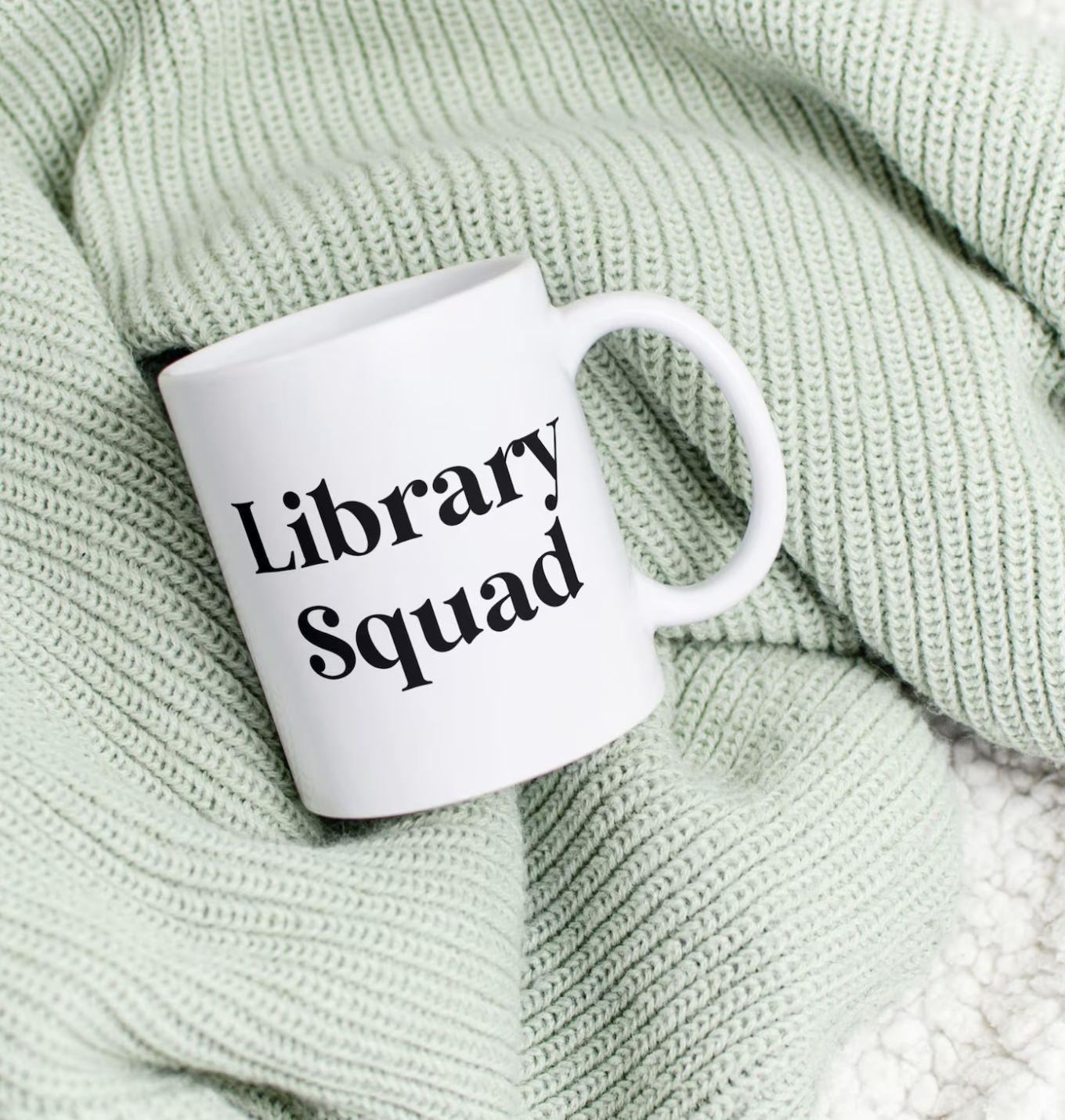 A white coffee mug with black text reading "library squad" laid against a pastel green knit blanket