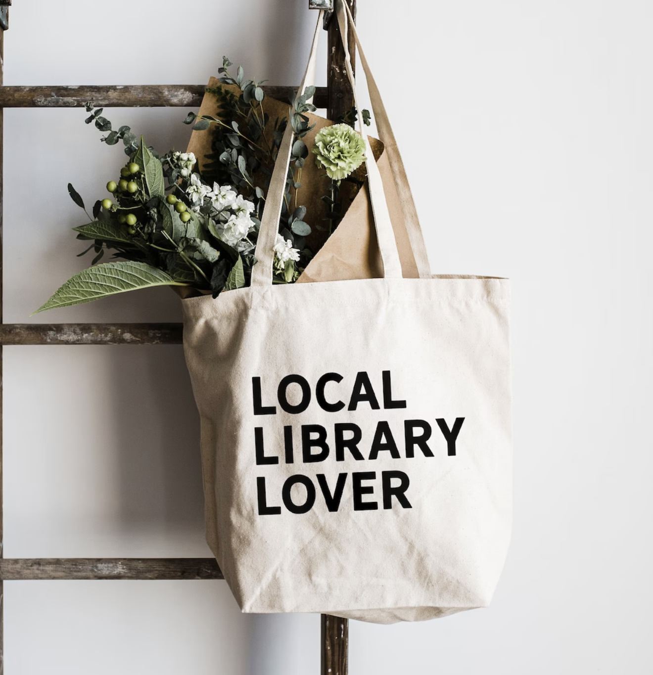 White canvas tote bag filled with fresh flowers with the saying "LOCAL LIBRARY LOVER" in black print across the front. 