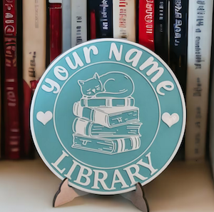 a round plaque for a bookshelf that has a cat on a stack of books illustration and can have your name customized to say it's your library