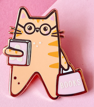 enamel pin of illustrated orange cat hugging a pink book and holding a pink purse that says "Books"