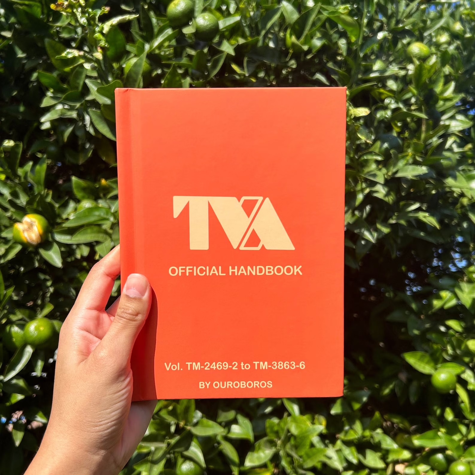 A hand holds up an orange journal with the words "TVA Official Handbook" written in gold in the center. At the bottom, also in gold, are volume numbers and the words "by Ouroboros."