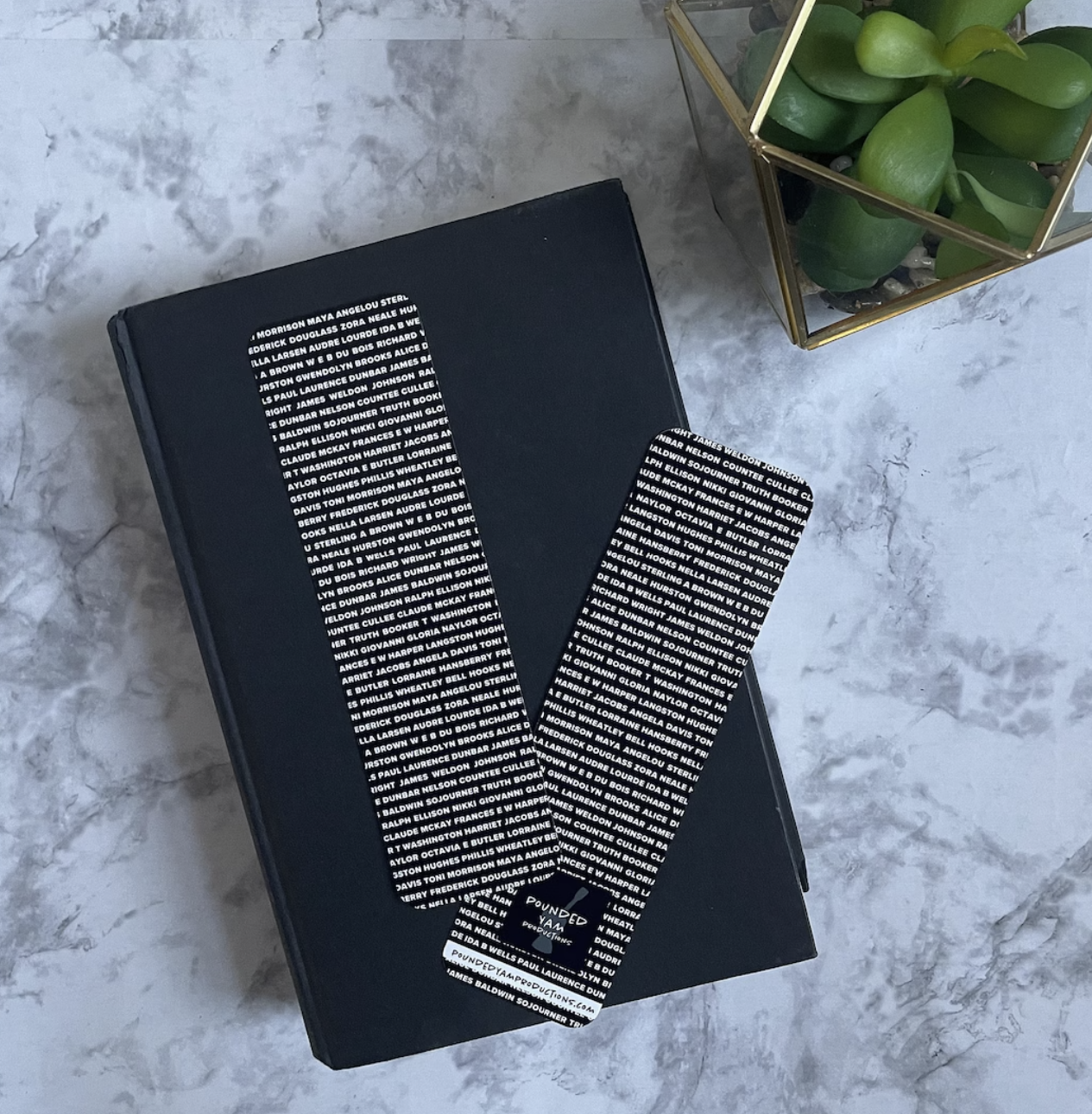 Picture of two black bookmarks covered in small white text listing Black authors like Toni Morrison and Maya Angleou set on a black book against a marble background