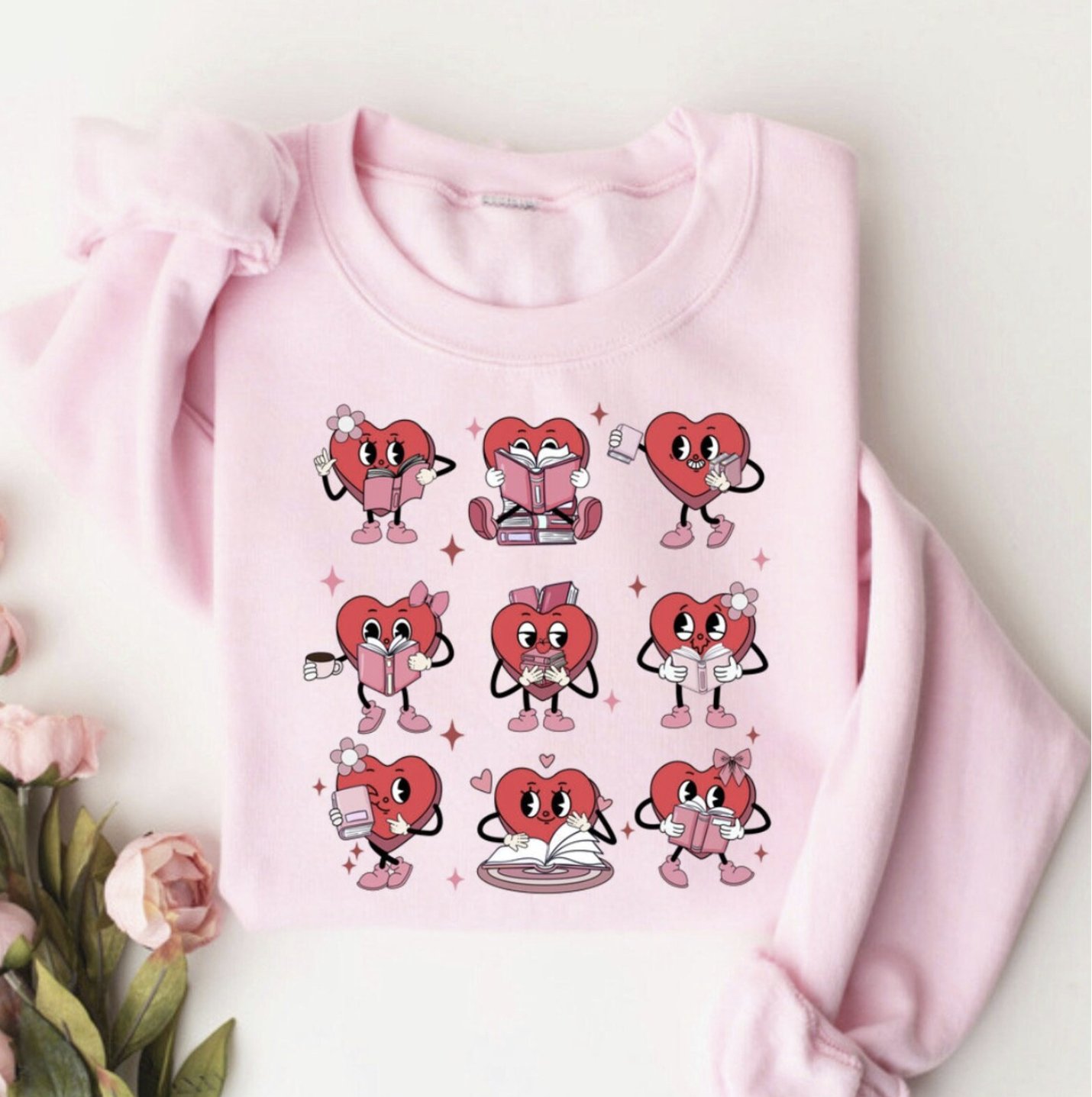 Pink crew neck sweatshirt with a grid of six red cartoon hearts reading books in various cute poses.