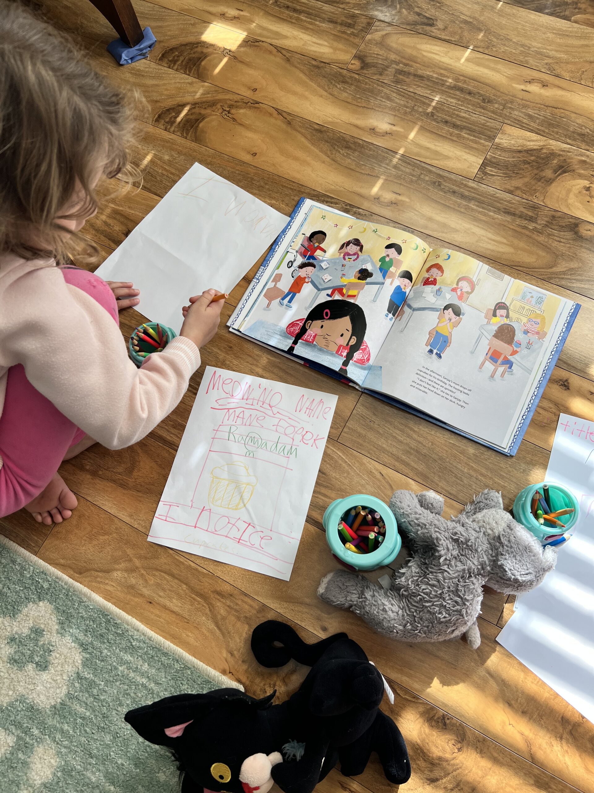 a kid draws on a piece of paper with a picture book open beside her and stuffed animals around her