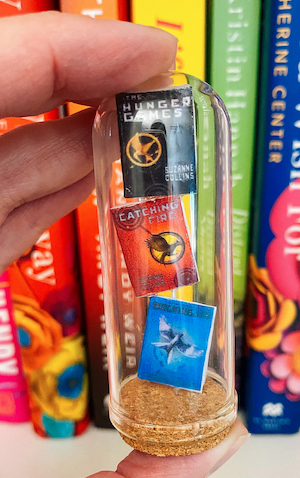 a glass tube on a cork stand with mini books of the Hunger Games trilogy stacked inside