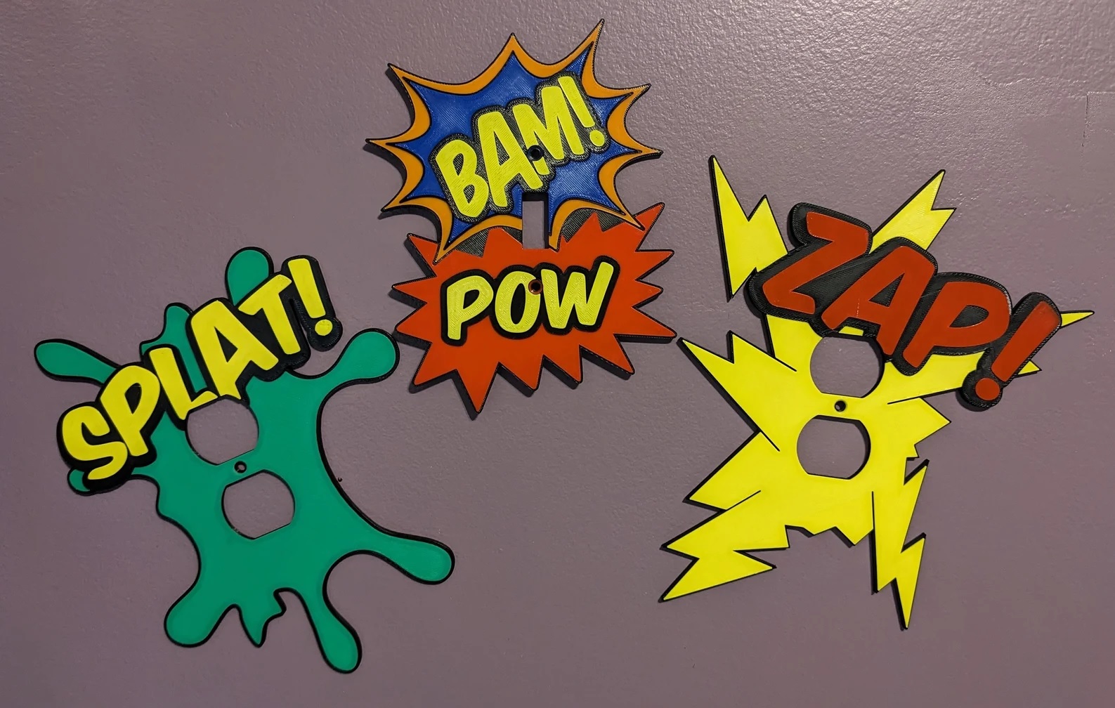 A set of three outlet covers, each resembling a brightly colored onomatopoeia from a comic book 