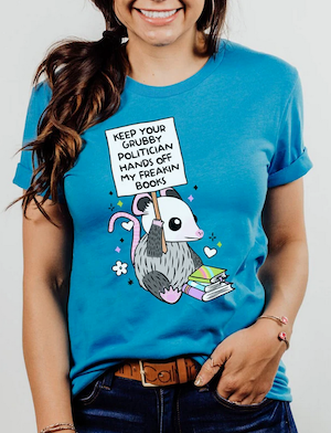 bllue tshirt with an illustrated possum holding a sign that says Keep Your Grubby Politician Hands Off My Books