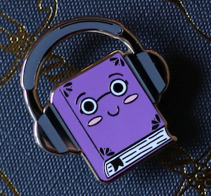 hard enamel pin of a purple book with adorable face wearing headphones