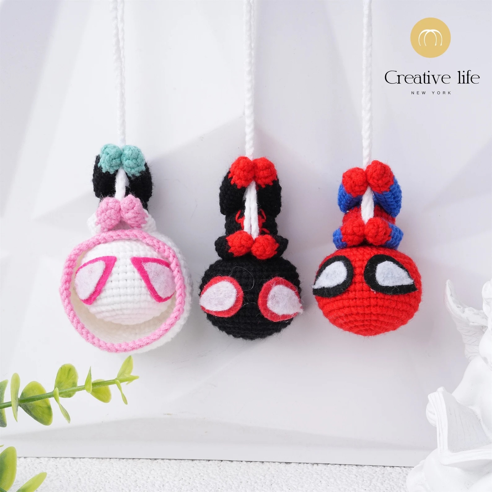 Small crocheted figures of Spider-Gwen, Miles Morales, and Peter Parker hanging upside-down from crocheted ropes