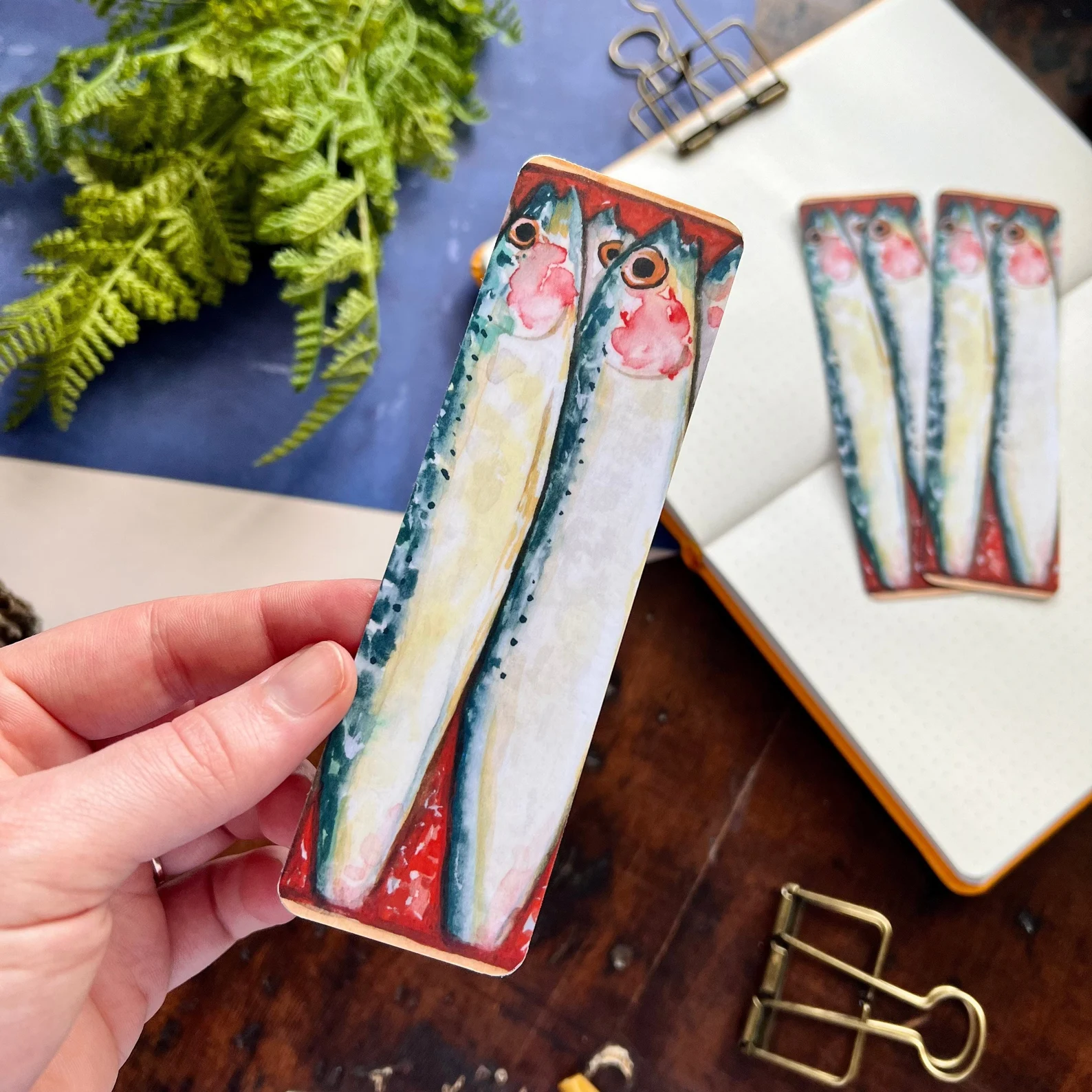 a photo of a bookmark that has an illustration of to sardines. The bookmark look like you are peering into their little sardine can