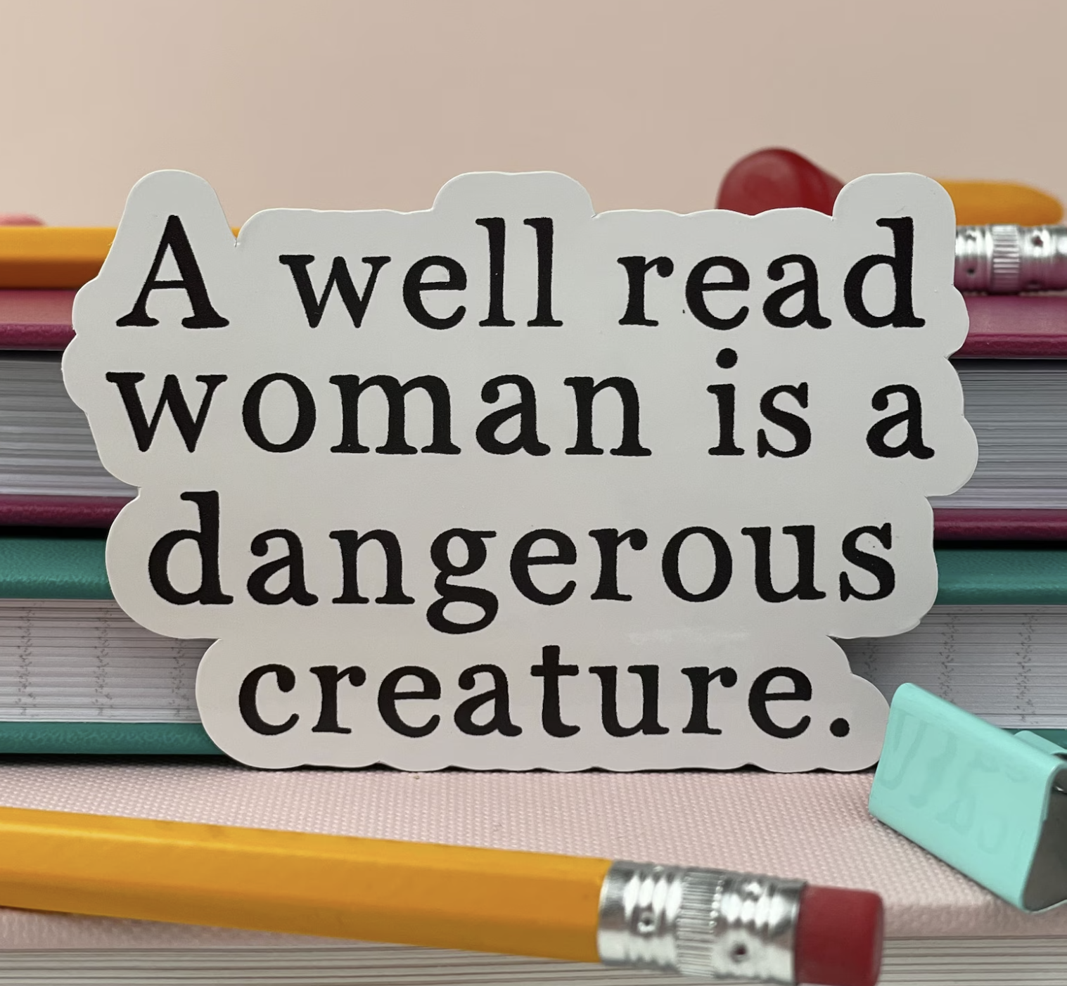 a white siker of black text that reads "a well read woman is a dangerous creature." The sticker rests against a stack of books.