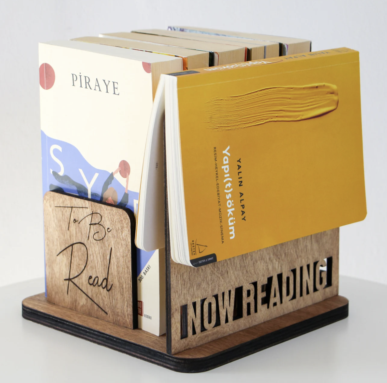 A small wooden square bedside book holder with a laser-cut front section that says "NOW READING" and room to hold five paperback books in a neat row.