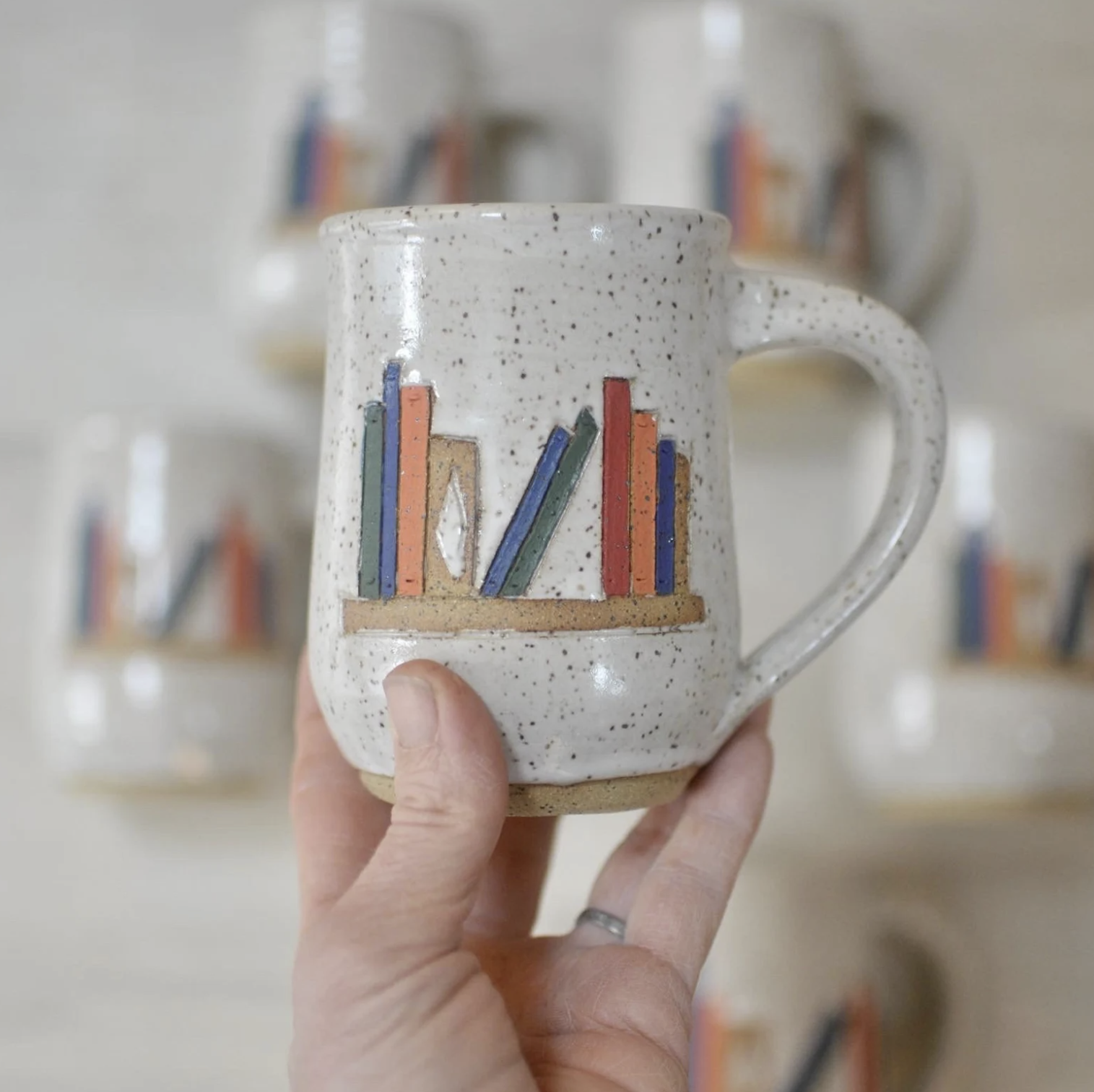 A handmade ceramic mug in a white speckled color with a curvy handle is held up by a white person's hand. The mug features an illustration of a single shelf with books in blue, green, orange, red, and tan colors. 