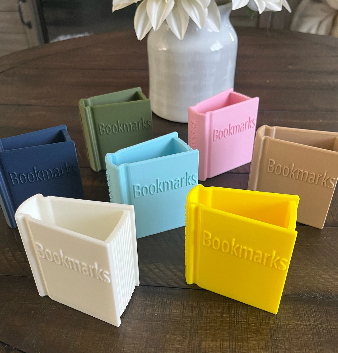 Image of an array of 3D printed book shaped containers in various colors embossed with the word "bookmarks" on the cover and spine and featuring an open top to hold bookmarks.