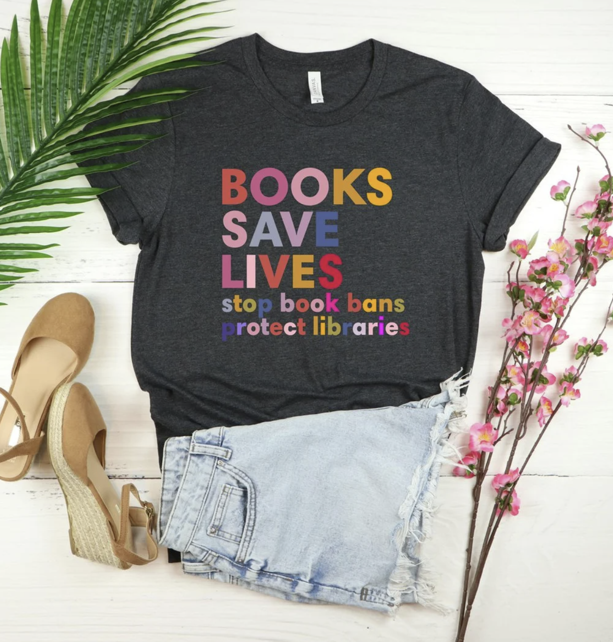 A charcoal colored tee-shirt against a white background and surrounded by a palm front, wedges, and flowers, proclaims in a colorful, fold font "BOOKS SAVE LIVES: stop book bans, protect libraries."