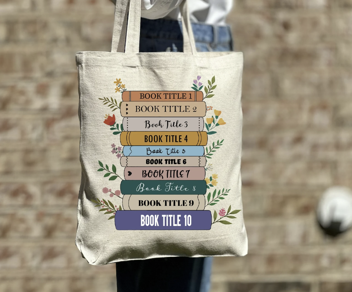 The lower body of a woman in a white shirt and jeans is visible holding up a canvas tote bag with a colorful book stack waiting to be customized with your favorite book titles.  