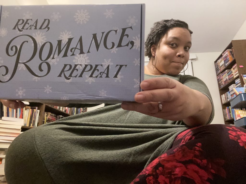 Photograph of Jess sitting crosslegged, holding up a pale blue and snowflake box around the size of a shoebox that says "Read, Romance, Repeat"