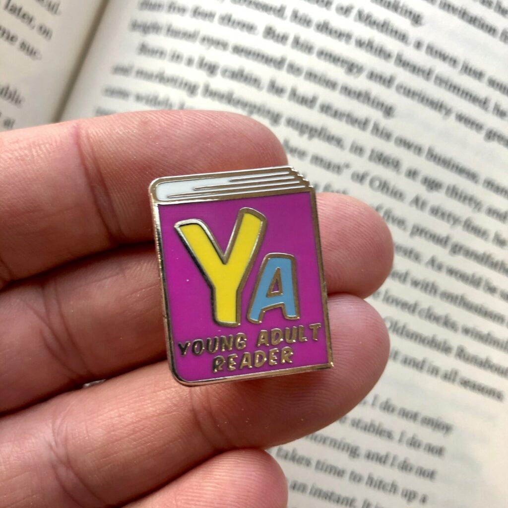 Image of purple enamel pin that reads "YA Young Adult Readerr"