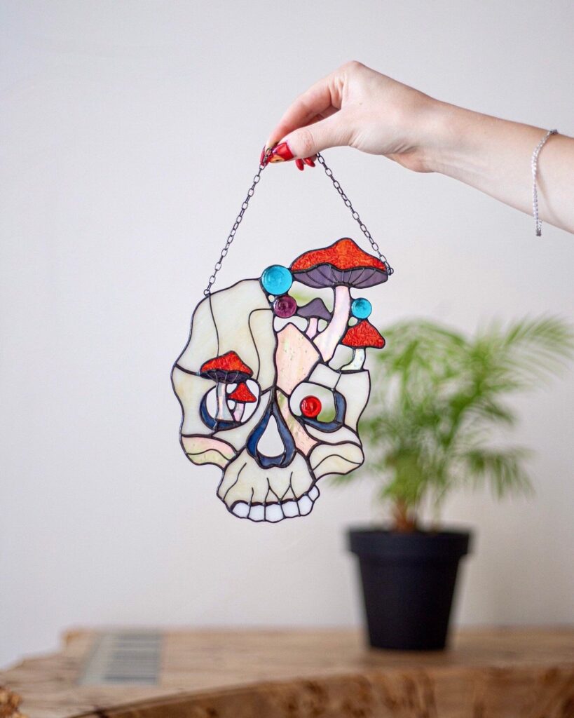 Stained glass suncatcher of a skull with fly amanita mushrooms growing out of the bone.