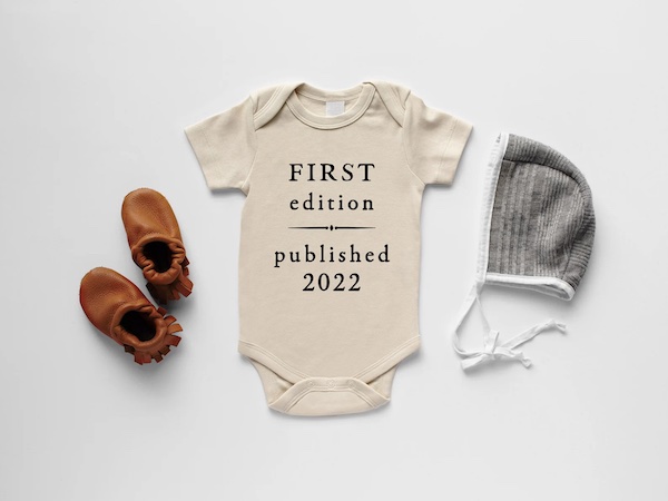 a cream colored baby onesie that says "FIRST edition --- published 2022"; there are also brown moccasin style booties and a gray cap