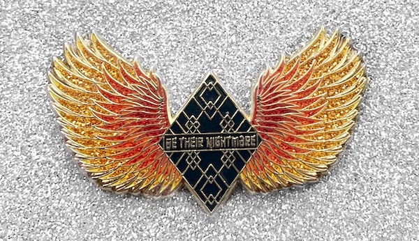 A pin that says "Be their nightmare" and has glittering, firey wings on either side of it.