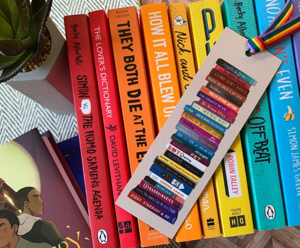 Image of a rainbow of queer YA books on a cream bookmark with a rainbow tassel. It is on top of a stack of queer YA books arranged in a rainbow color scheme. 