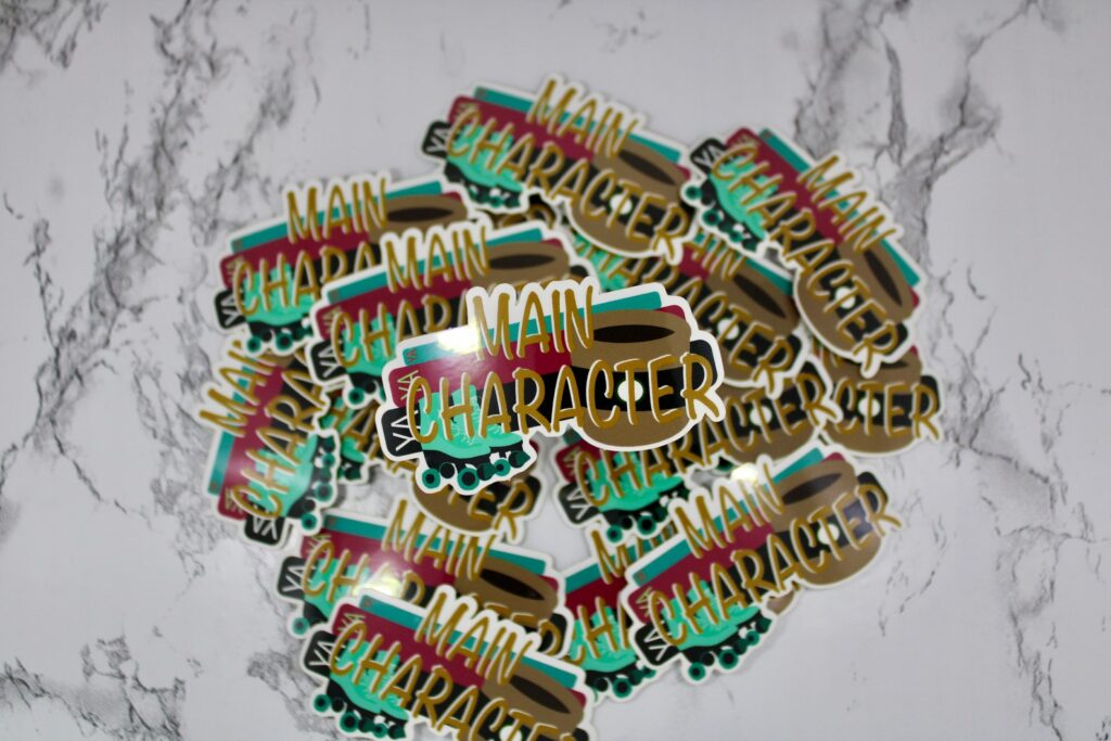 Pile of stickers that read "main character energy" in light brown. The background colors on the sticker are green, maroon, black, and light brown. 