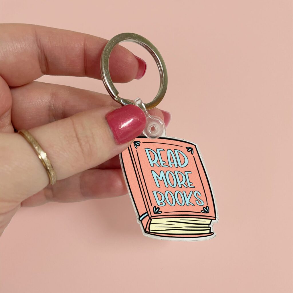 image of a peach colored keychain being held by a white hand in front of a light pink background. The keychain reads "read book books."