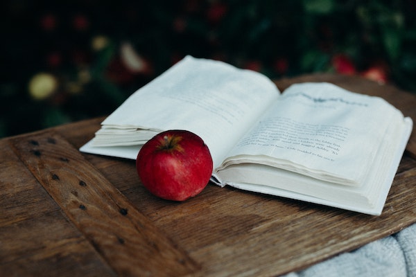 a photo of a book with some slight water damage on the pages lying open, with a red apple next to it. they are on a wooden table.