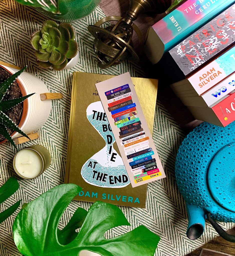 Image of a colorful bookmark made up of YA book covers. It's sitting on top of a cover of Adam Silvera's They Both Die at the End. 