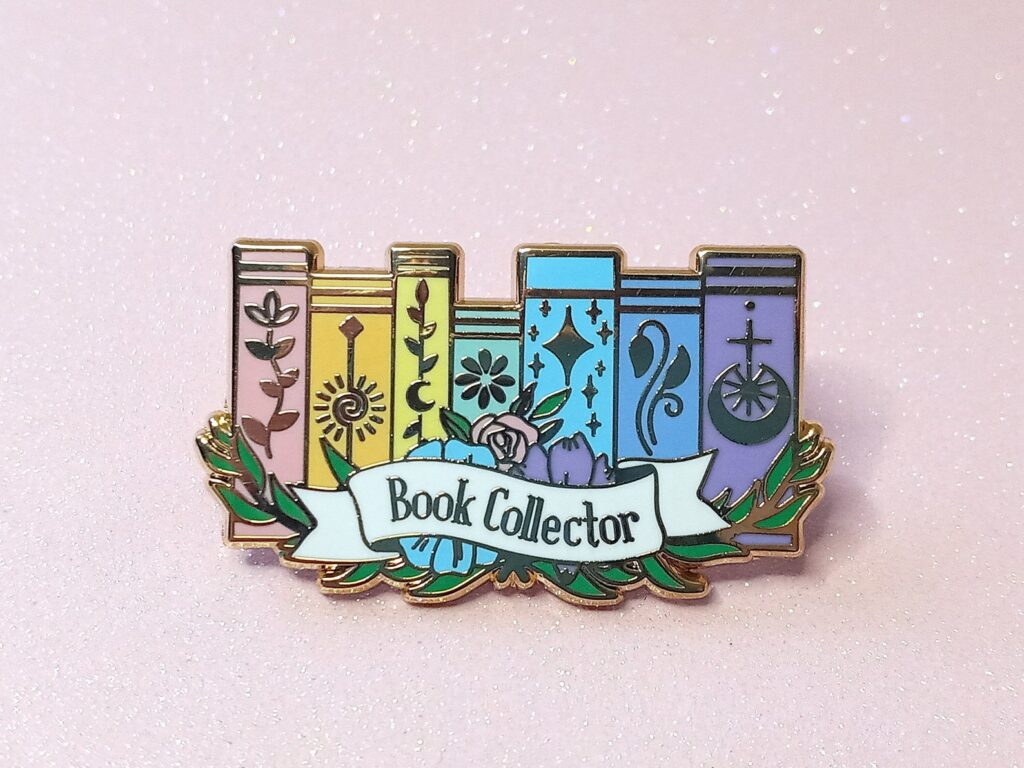 Image of an enamel pin on a pink background. The pin has seven book spines in a rainbow of colors. The text on the pin reads "book collector."