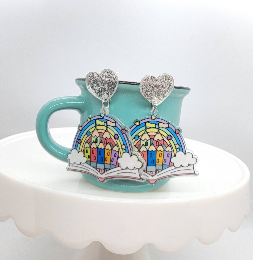Bright colored earrings. They are dangling on a teal cup. The earrings each feature an open book with a castle and rainbow emerging from it. 