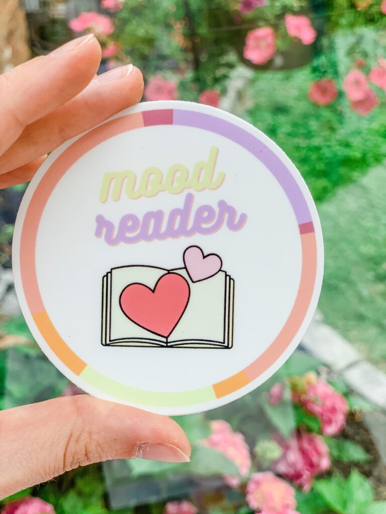 Image of a round sticker. It's pink, peach, and purple. In the center it reads "Moo Reader" with a book that has hearts coming out of it. 