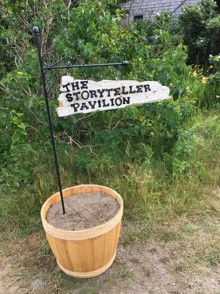 a pot of dirt with a black metal sign sticking out that reads "The Storyteller Pavilion"