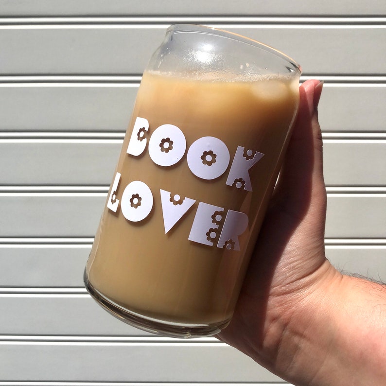 Image of a glass being held by a light hand. The glass has white font reading "book lover."