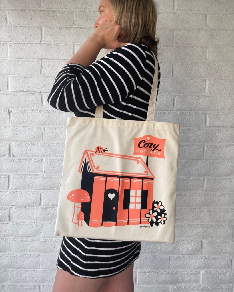 Image of a white person in navy and white stripes.  They're wearing a canvas tote back that has a pink and black image on it of a house made of book spines. It reads 'cozy cottage."