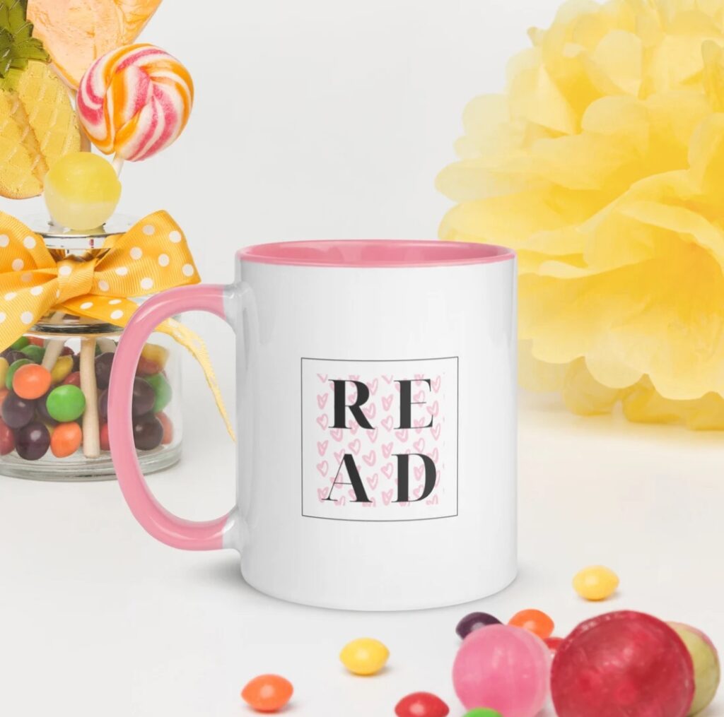 Image of a white mug with a pink handle and pink interior. The mug has a black box with the letters R E A D inside. 
