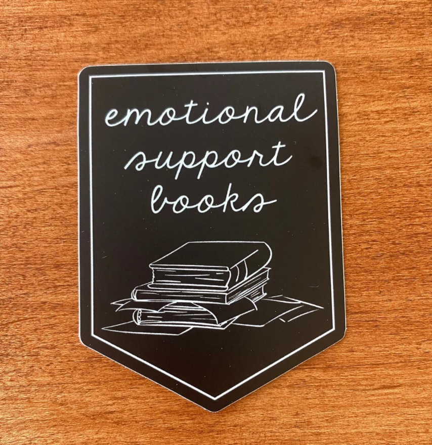 Image of a black pennant shaped sticker. It has white text reading "emotional support book," with a small sketch of white books.