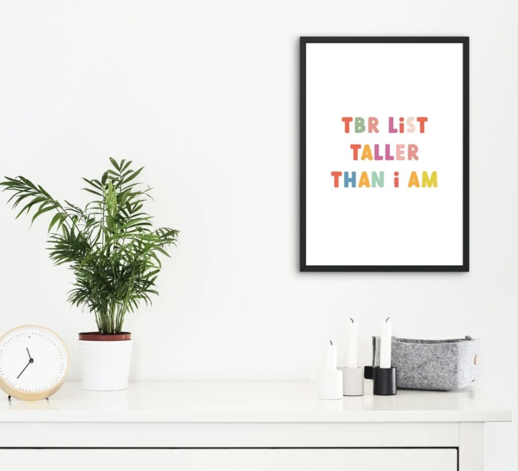 Image of a colorful print in a simple black frame hanging on a white wall. The print reads "tbr list taller than i am."