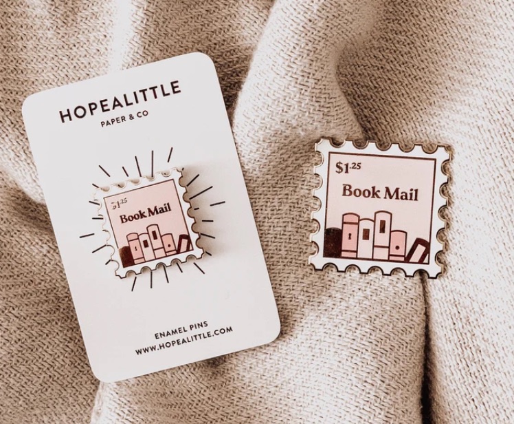 Image of the same enamel pin two times. It looks like a postage stamp and says "book mail $1.25." There are book spines on it, and the colors are pastel peaches and pinks. 