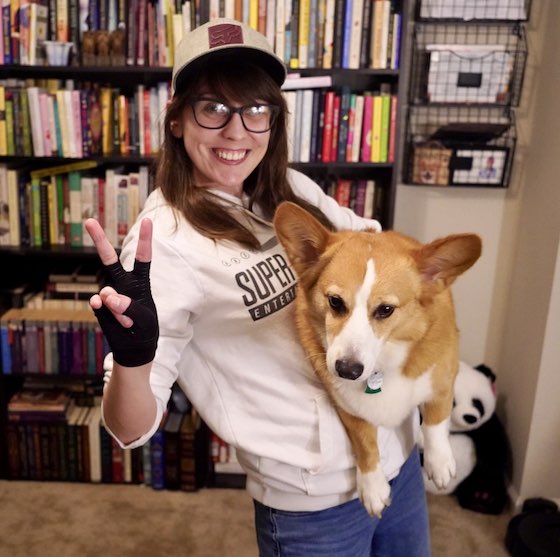 A photo of Kendra, a white woman with brunette hair. She wearing a white sweatshirt and grey dad hat. She's holding her red and white Pembroke Welsh Corgi, Dylan. Dylan is not thrilled to be having his photo taken.